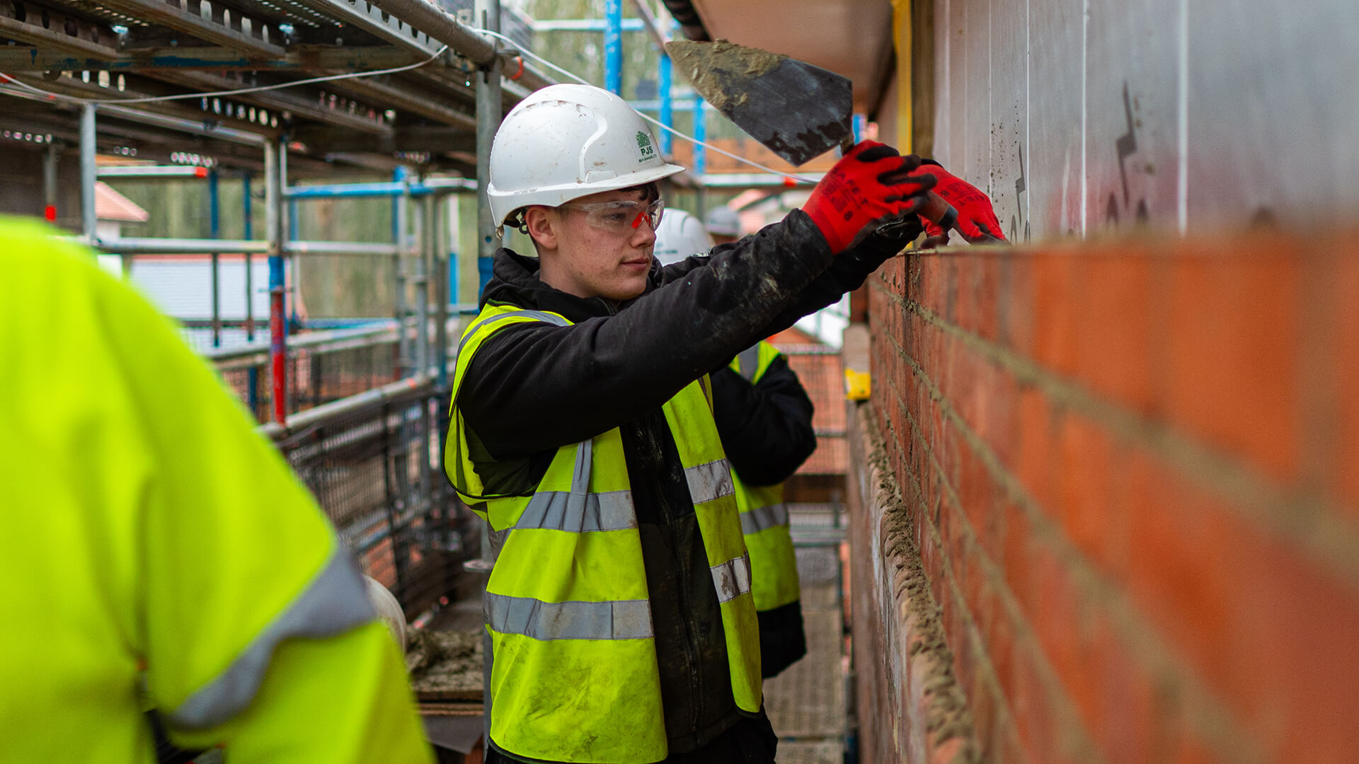 Student wearing hard hat and hi-vis vest smiling at camera and leaning against brick wall.