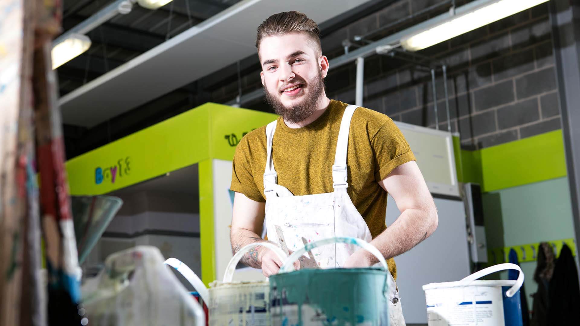 A young man smiling whilst wearing white overalls that are splattered in paint and standing next to a pile of paint tins.