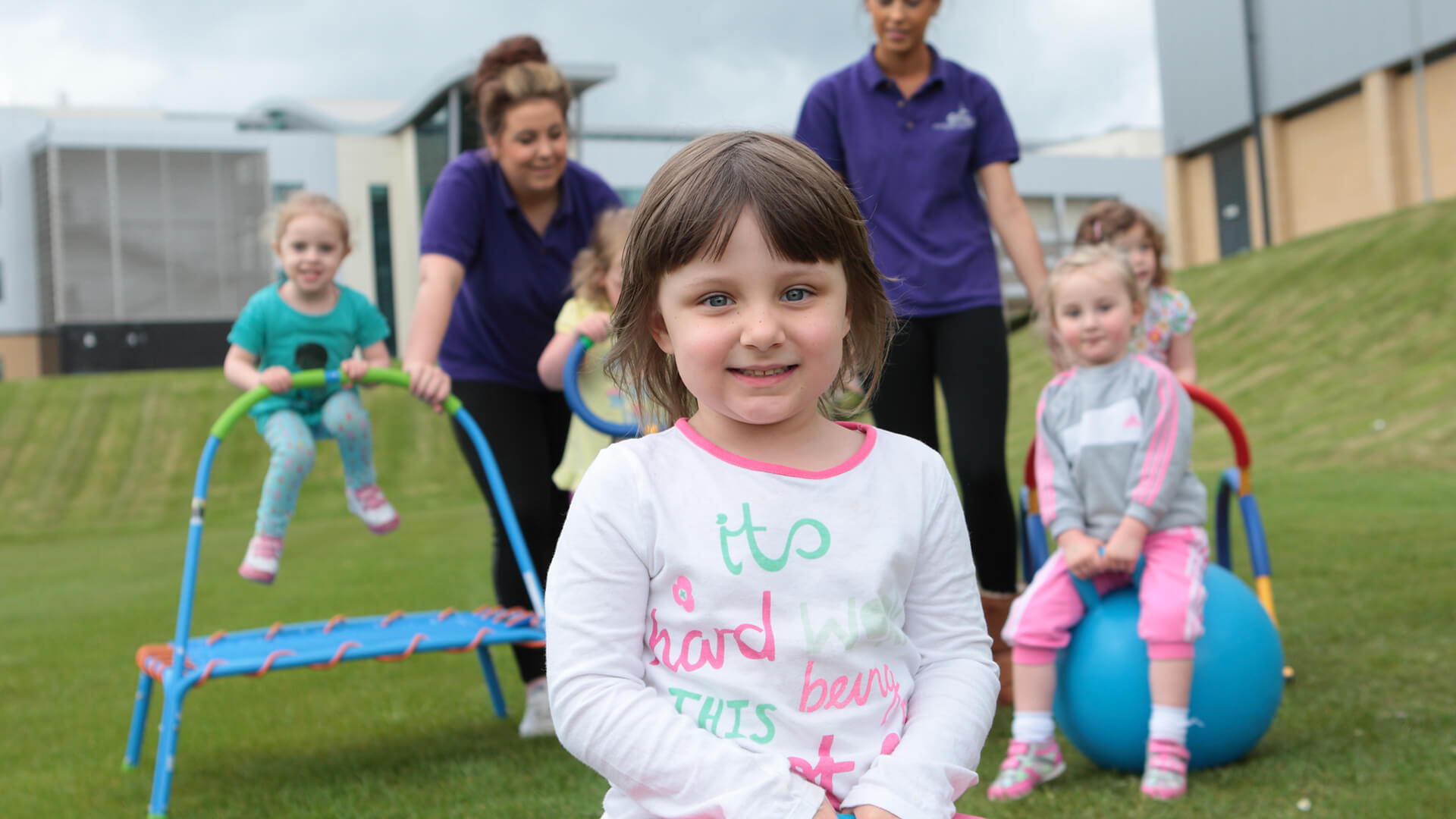 Five small children and two nursery nurses playing outside on a patch of grass.