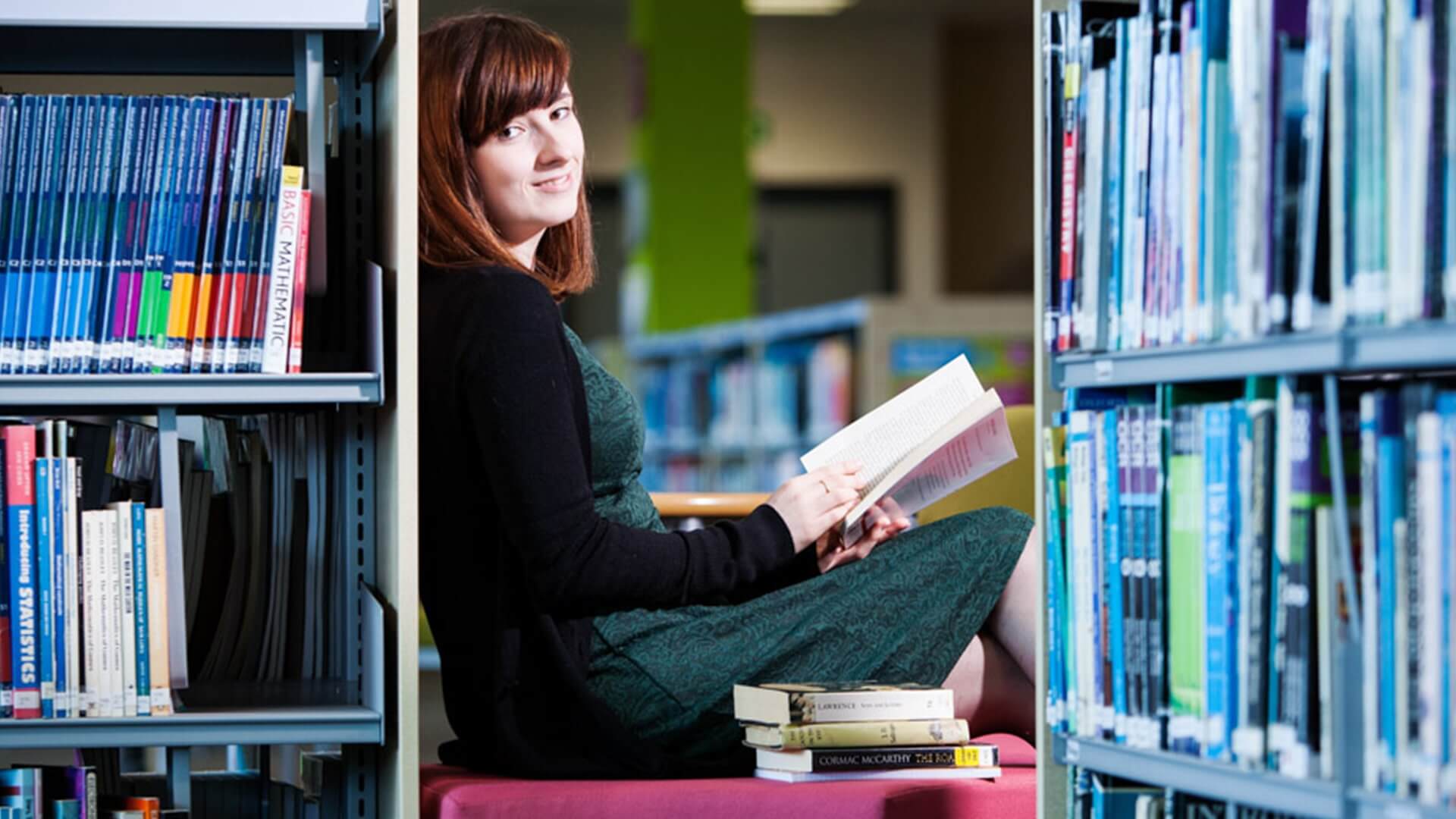 A young woman sat in a library, reading a book and smiling.