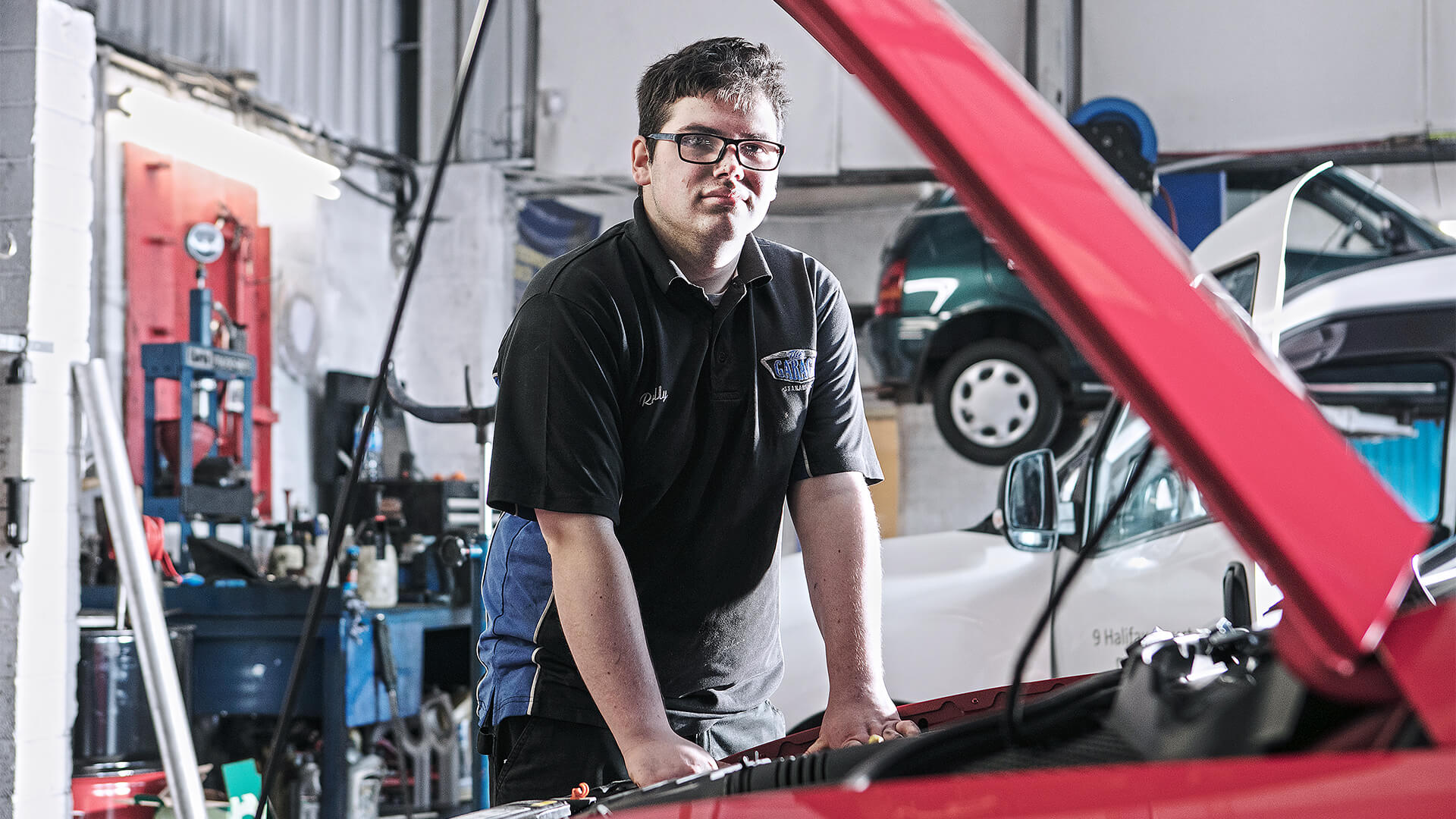 Man standing next to a car with the bonnet up, in a garage full of other cars and tools.