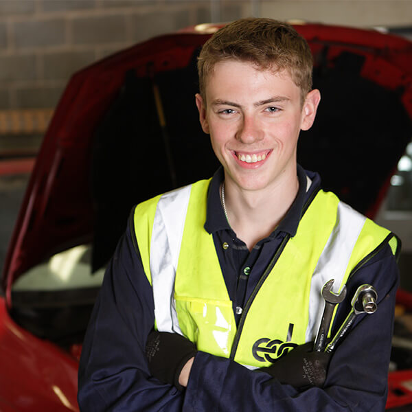 A young man smiling in a high-vis jacket whilst standing in front of a car.