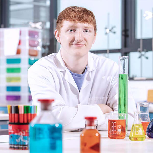 Young man in a white lab coat sat at a desk that has many beakers with coloured liquids on it.