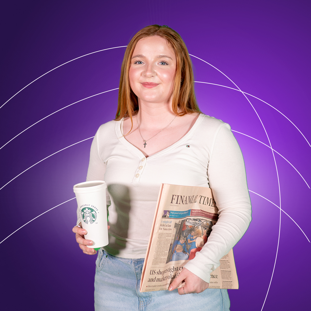 Young women holding starbucks cup in one hand and financial times under her arm smiling at camera on a purple background.