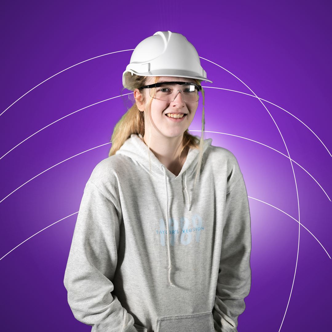 Young women wearing hard hat and safety glasses smiling at camera on a purple background.