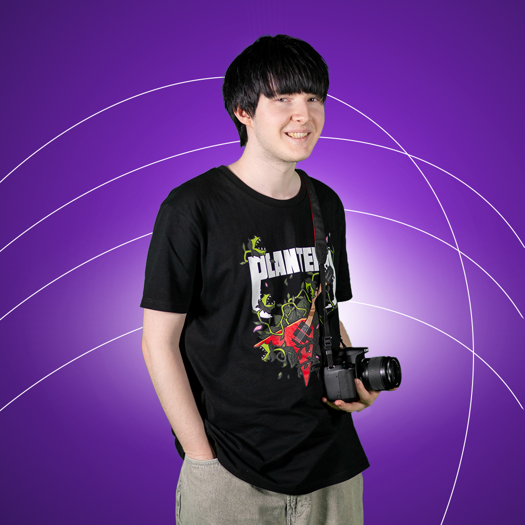 Young man holding digital camera smiling at camera on a purple background.