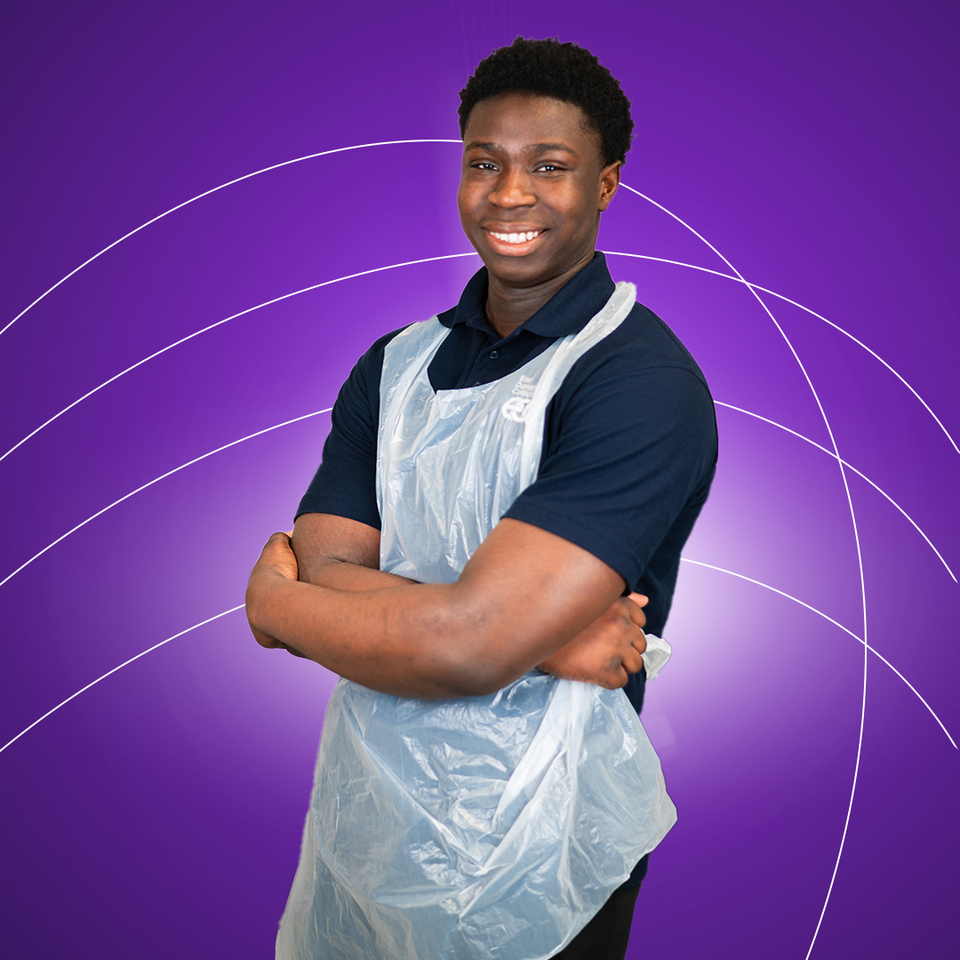 Young man wearing medical apron standing with arms crossed and smiling at camera on a purple background.