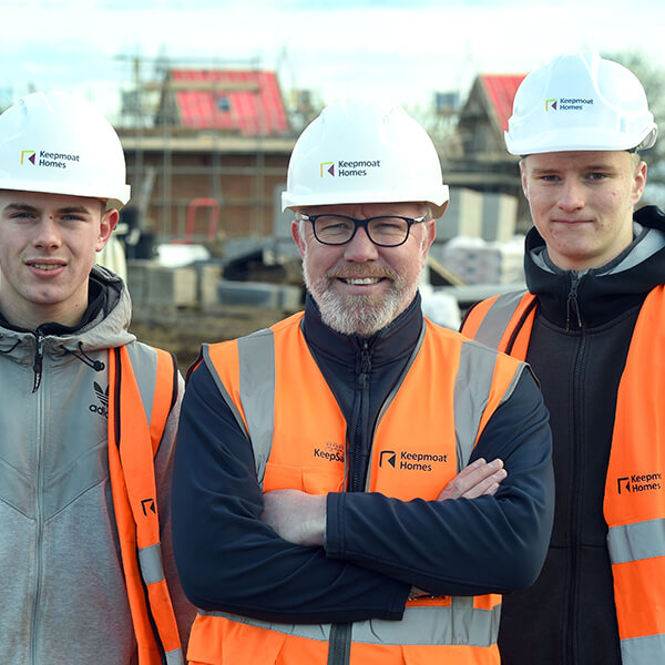 Three men in high-vis jackets and white hard hats standing in front of a house development site.