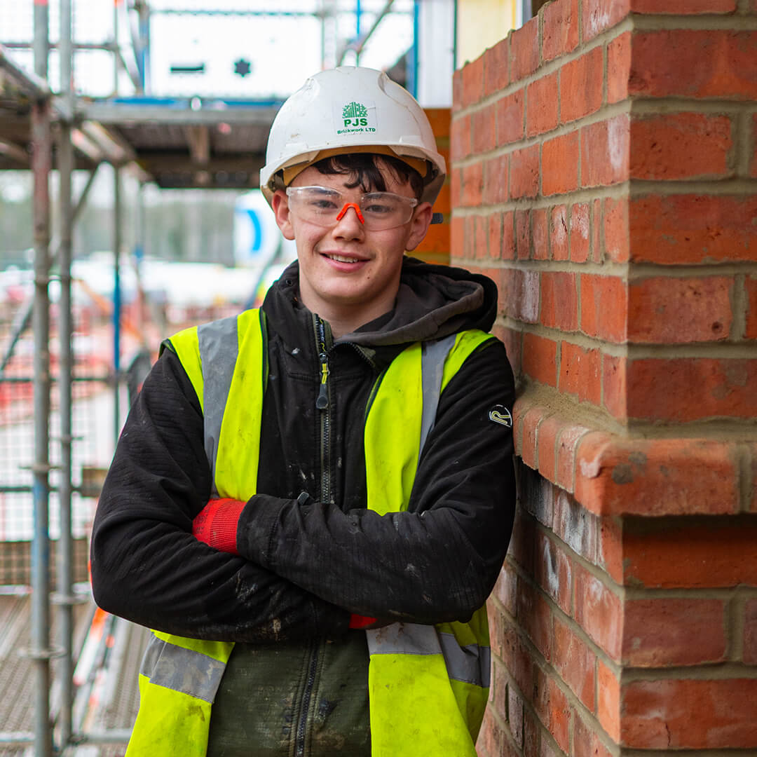 Student wearing hard hat and hi-vis vest smiling at camera and leaning against brick wall.