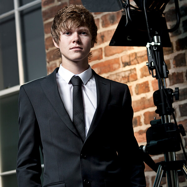 Young man in a smart black suit in front of a brickwork.