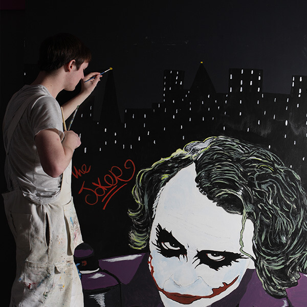 A student paying a mural on the wall of The Joker from Batman.