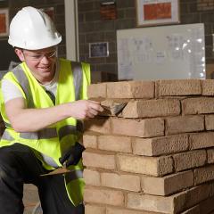 A bricklaying student in a white hard hat holding a trowel kneeling nest to a stack of bricks
