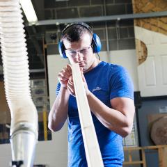 A man wearing a blue t-shirt and ear protectors, looking down a long length of cut timber