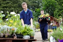 Two young male students in a garden centre, one pushing a trolley of plants, the other carrying plants