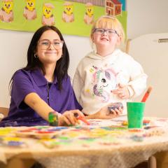 Smiling student in purple top with small child playing with paint
